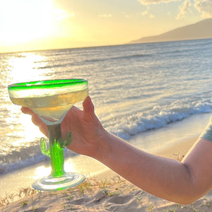 A hand holding a Aunty Lei's MauiRita in a cactus margarita glass in the beach at sunset.
